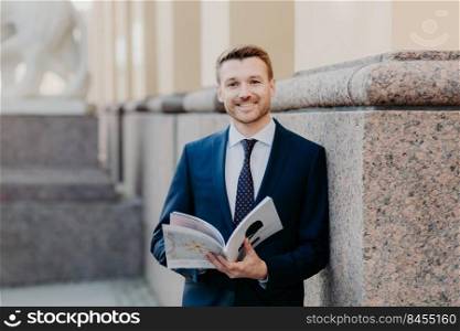 Prosperous successful businessman in formal outfit holds journal, has happy expression, smiles gladfully, enjoys dinner break after hard working day, being in good mood after business meeting