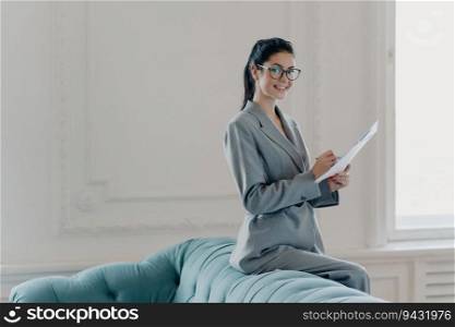 Prosperous woman financial manager studies documents in spacious home office, dressed elegantly, writing information, looks pleased at the camera. Paperwork concept.