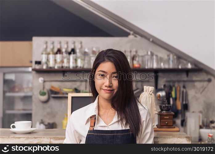 Protrait of young Asian female barista smiling to welcome her customer to coffee shop with a background of beverage bar counter. Young female barista smile in her coffee shop. Food and drink concept.