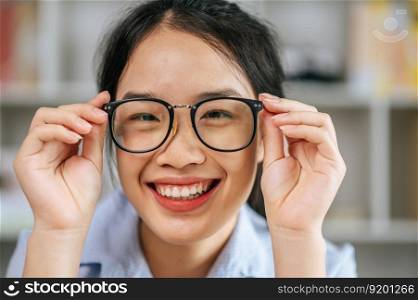 Protrait young asian woman happy positive smile and use hand touch eyeglasses and looking at camera at living room