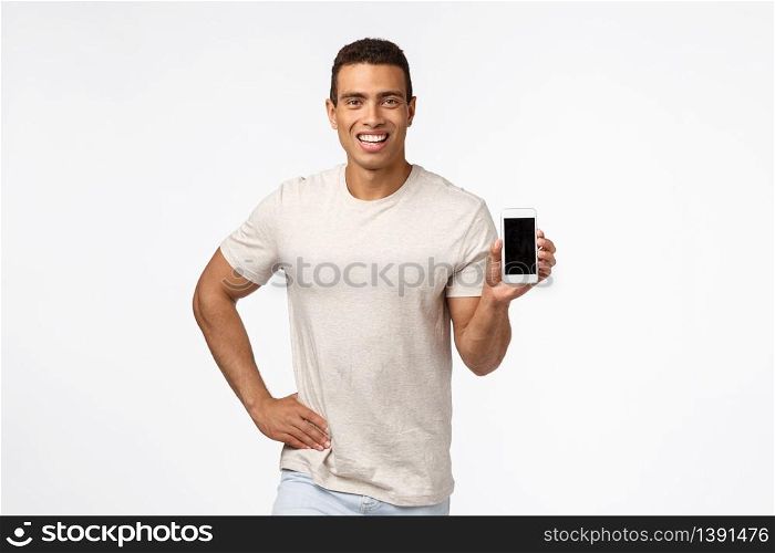 Proud and assertive handsome hispanic man in t-shirt, holding smartphone, promote application or shopping site, smiling delighted, give recommendation, advertise gadget or app, white background.. Proud and assertive handsome hispanic man in t-shirt, holding smartphone, promote application or shopping site, smiling delighted, give recommendation, advertise gadget or app, white background