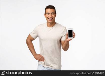 Proud and assertive handsome hispanic man in t-shirt, holding smartphone, promote application or shopping site, smiling delighted, give recommendation, advertise gadget or app, white background.. Proud and assertive handsome hispanic man in t-shirt, holding smartphone, promote application or shopping site, smiling delighted, give recommendation, advertise gadget or app, white background