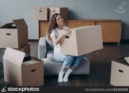 Proud european girl relocates to new home. Happy young woman moves alone. Lady is sitting in armchair among packed cardboard boxes. Independence, life choice and success concept.. Proud european girl relocates to new home. Happy young woman moves alone.