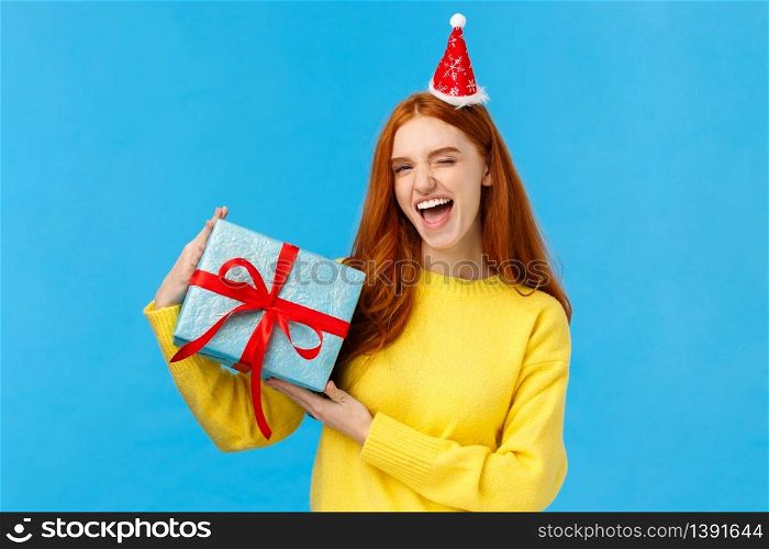 Proud girl wrapped gift for girlfriend herself, winking sassy and pleased, smiling joyfully, wearing christmas hat, holding box with present and congratulating friend happy holidays, bllue background.. Proud girl wrapped gift for girlfriend herself, winking sassy and pleased, smiling joyfully, wearing christmas hat, holding box with present and congratulating friend happy holidays, bllue background