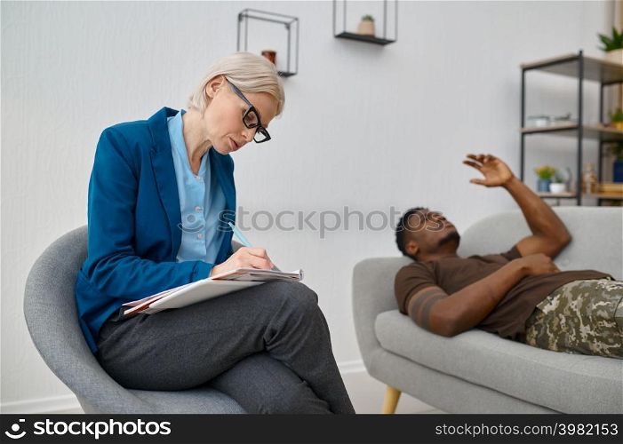 Psychologist making notes on therapy session with sad soldier. War veteran talking lying on couch. Psychologist making notes while male soldier talking
