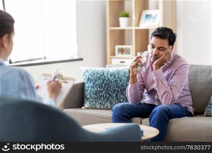 psychology, mental therapy and people concept - sad young indian man patient with glass of water and woman psychologist at psychotherapy session. man and psychologist at psychotherapy session