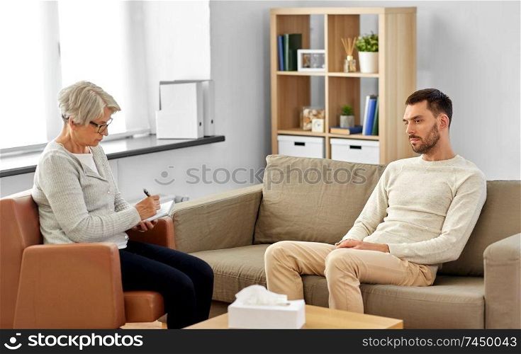 psychology, mental therapy and people concept - senior woman psychologist taking notes to notebook and sad young man patient at psychotherapy session. senior woman psychologist and sad man patient