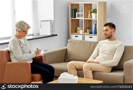 psychology, mental therapy and people concept - senior woman psychologist taking notes to notebook and sad young man patient at psychotherapy session. senior woman psychologist and sad man patient