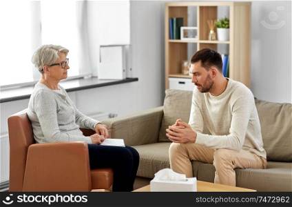 psychology, mental therapy and people concept - senior woman psychologist talking to sad young man patient at psychotherapy session. senior woman psychologist and young man patient
