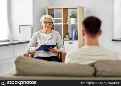 psychology, mental therapy and people concept - senior woman psychologist with clipboard and young man patient at psychotherapy session. senior woman psychologist and young man patient