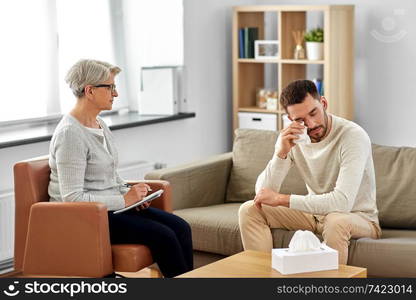 psychology, mental therapy and problem concept - senior woman psychologist taking notes to notebook and sad young man patient crying and wiping eyes at psychotherapy session. senior woman psychologist and crying man patient