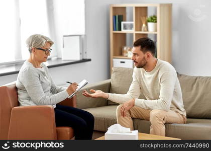 psychology, mental therapy and problem concept - senior woman psychologist taking notes to notebook and sad young man patient at psychotherapy session. senior woman psychologist and sad man patient