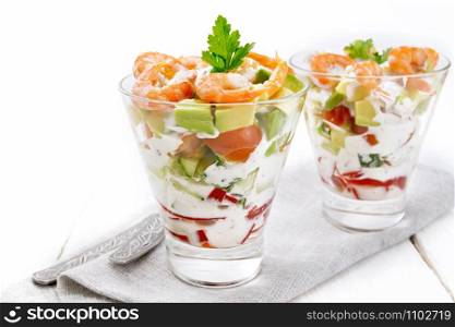 Puff salad with shrimp, avocado, fresh cucumber, sweet pepper and tomato, seasoned with yogurt sauce in two glass glasses on a napkin, bread and forks on a white wooden board background