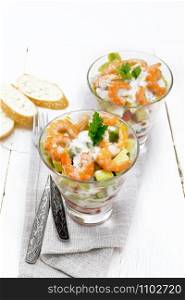 Puff salad with shrimp, avocado, fresh cucumber, sweet pepper and tomato, seasoned with yogurt sauce in two glass glasses on a towel, bread and forks on a light wooden board background