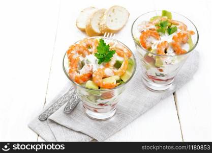 Puff salad with shrimp, avocado, fresh cucumber, sweet pepper and tomato, seasoned with yogurt sauce in two glass glasses on a towel, bread and forks on a white wooden board background