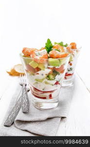 Puff salad with shrimp, avocado, fresh cucumber, sweet pepper and tomato, seasoned with yogurt sauce in two glass glasses on a napkin, bread and forks on a wooden board background