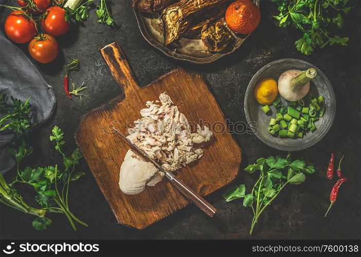 Pulled chicken breast on cutting board with knife with vegetables ingredients on dark rustic background. Top view. Healthy food eating. Low carb fitness dieting. Easy cooking