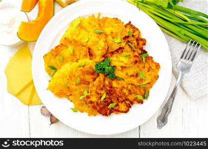 Pumpkin and cheese fritters with ginger in a plate, sour cream in a bowl, napkin, parsley and fork on background of light wooden board from above