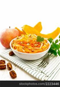 Pumpkin, carrot and apple salad with pecans seasoned with vegetable oil in a bowl on a towel, mint on light wooden board background