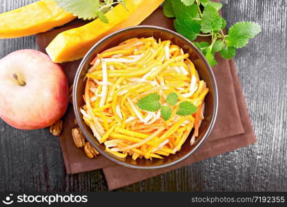 Pumpkin, carrot and apple salad with pecans seasoned with vegetable oil in a bowl on a napkin, mint on dark wooden board background from above