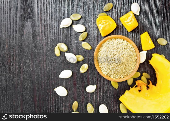 Pumpkin flour in a bowl, seeds on the table, slices of vegetable on dark wooden board background from above
