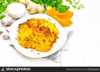 Pumpkin fritters in a white plate on napkin, sour cream in a bowl, parsley, garlic and ginger on wooden board background. Pancakes of pumpkin in plate on light board