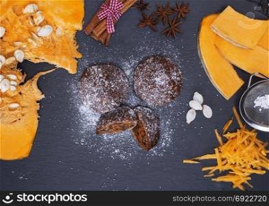 pumpkin muffins sprinkled with powdered sugar on a black background and pieces of fresh pumpkin, top view