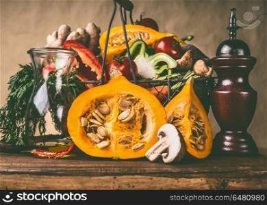 Pumpkin on kitchen table with basket of vegetables ingredients at rustic wall background, front view. Healthy vegetarian food and eating concept. Autumn seasonal eating