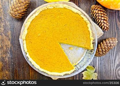 Pumpkin pie in a glass pan on sackcloth, cones and maple leaf on the background of the wooden planks on top