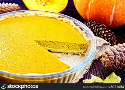 Pumpkin pie in a glass pan on sackcloth, pumpkins, pine cones and maple leaf on the background of dark wood planks