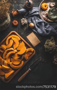 Pumpkin slices on baking sheet on dark rustic kitchen table background with knife and ingredients, top view. Copy space