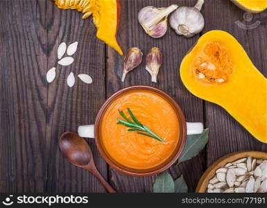 pumpkin soup in a ceramic plate with a wooden spoon on a brown table, next to pumpkin seeds and garlic, empty space on the left