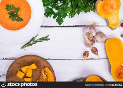 pumpkin soup in a white plate and ingredients for cooking on a white wooden background, empty space in the middle, top view