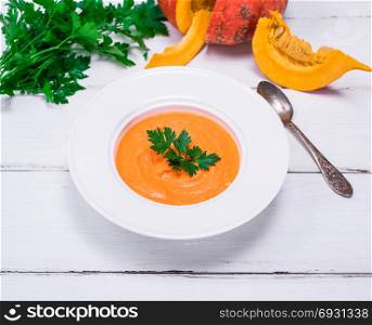 Pumpkin soup puree in a round white plate and fresh pumpkin on a white wooden table, top view
