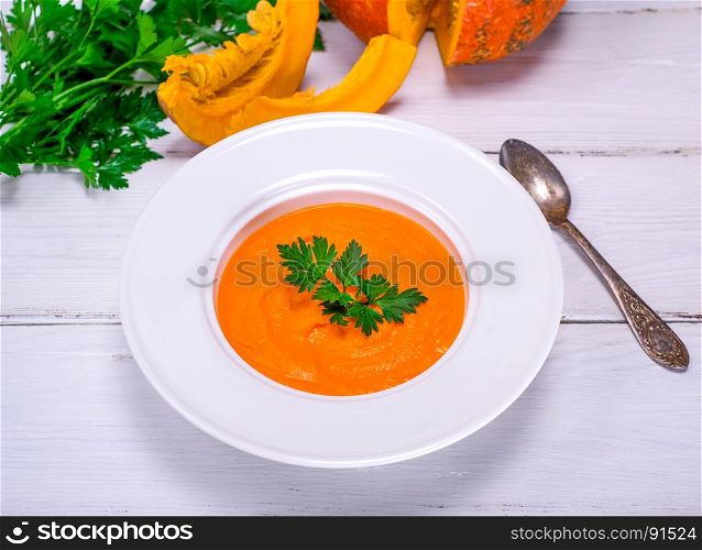 Pumpkin soup puree in a round white plate and fresh pumpkin on a white wooden table