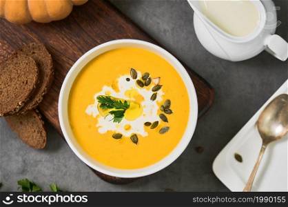 Pumpkin soup with parsley, cream and pumpkin seeds in a white bowl, top view. Layout on gray background with ingredients and bread