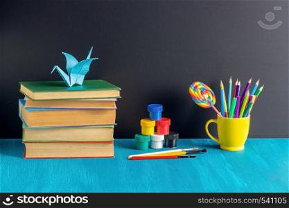 Pupils workplace. Books stationery paint gouache and origami crane on blue table on background black chalkboard Concept Education and Back to school.. Pupils workplace. Books stationery paint gouache and origami crane on blue table on background black chalkboard Concept Education and Back to school