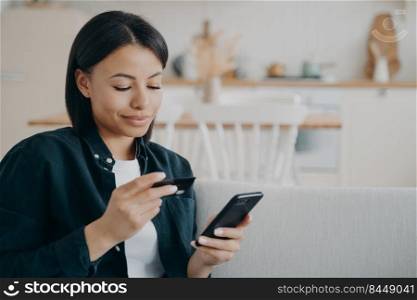 Purchasing online from home on quarantine. Young happy european woman is holding smartphone and credit card sitting on couch. Spanish girl ordering through internet. Money transfer and e-commerce.. Purchasing online from home on quarantine. Spanish girl ordering through internet.
