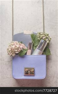 Purple handbag with hydrangea flowers and cosmetic products on pale grey concrete table. Lifestyle concept. Top view.