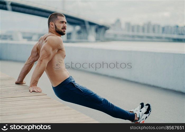 Purposeful bearded adult man trains biceps while doing reverse push up exercises poses with naked muscular torso concentrated away breathes fresh air, likes fitness and sport. Workout outside
