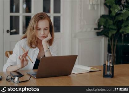 Puzzled female employee or student, sitting at work desk with laptop, studying or working online. Focused businesswoman, looking at computer screen, thinking of problem solution, making decision.. Focused female employee sitting at desk with laptop working online, thinking of problem. Remote job