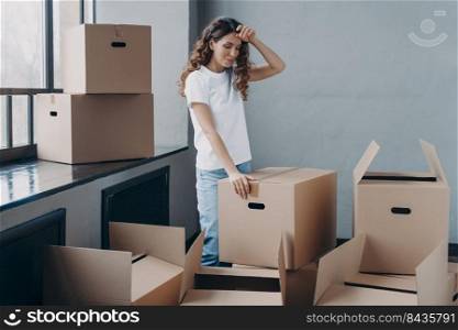 Puzzled woman unpacking cardboard boxes. Tired girl in new apartment. Lady touches forehead with hand, gesture of tired person. Packing things, shipping service and relocation concept.. Tired woman unpacking cardboard boxes. Packing things, shipping service and relocation concept.