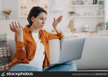 Puzzled young woman has online conference at home. European lady with laptop is talking at internet meeting. Confused girl is telling a problem. Businesswoman is working while quarantine.. Puzzled european woman with laptop is talking at internet meeting and telling a problem.