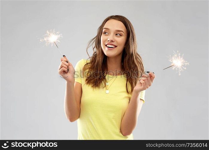 pyrotechnics and people concept - smiling young woman or teenage girl in blank yellow t-shirt with two sparklers over grey background. happy teenage girl with two sparklers