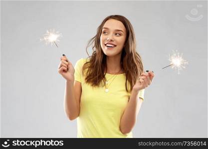 pyrotechnics and people concept - smiling young woman or teenage girl in blank yellow t-shirt with two sparklers over grey background. happy teenage girl with two sparklers