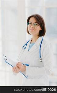 Qualified cardiac expert with stethoscope prepares for health seminar, gives consultancy about first aid, writes down information in clipboard, wears white gown, ready to give medic care help