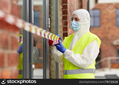 quarantine and pandemic concept - healthcare worker in protective gear or hazmat suit, medical mask, gloves and goggles enclosing building with caution tape outdoors. healthcare worker sealing door with caution tape