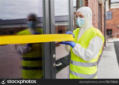 quarantine and pandemic concept - healthcare worker in protective gear or hazmat suit, medical mask, gloves and goggles enclosing building with caution tape outdoors. healthcare worker sealing door with caution tape