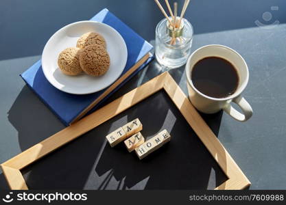 quarantine, epidemic and safety concept - close up of chalkboard with stay at home words on wooden toy blocks, coffee cup, cookies and book with aroma reed diffuser on windowsill. chalkboard with stay at home words on toy blocks