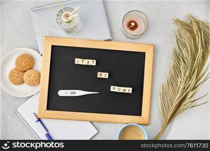 quarantine, epidemic and safety concept - close up of chalkboard with stay at home words on wooden toy blocks and thermometer, coffee cup, cookies and candle on table. chalkboard with stay at home words on toy blocks